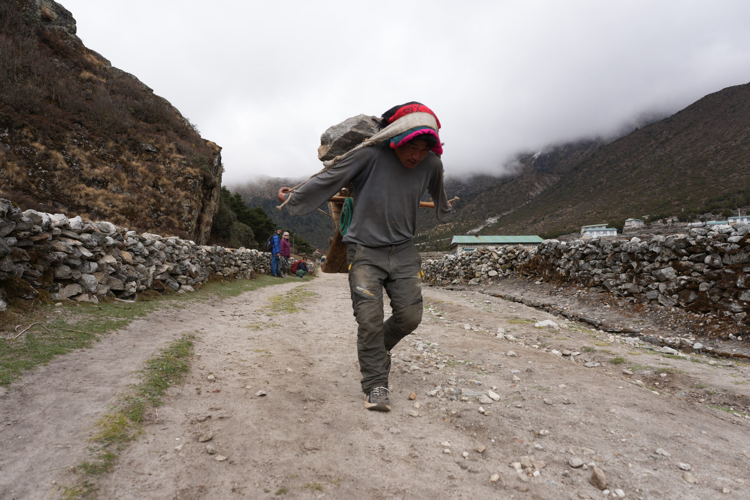A Sherpa laborer carries a large load of rocks in a woven back basket supported by a forehead strap as he walks along a dirt trail in the Everest region of Nepal. Stone walls line the path, while low clouds cling to the surrounding Himalayan mountains, highlighting the demanding conditions in which Sherpas build strength and endurance. Namche Bazaar, Nepal 2025