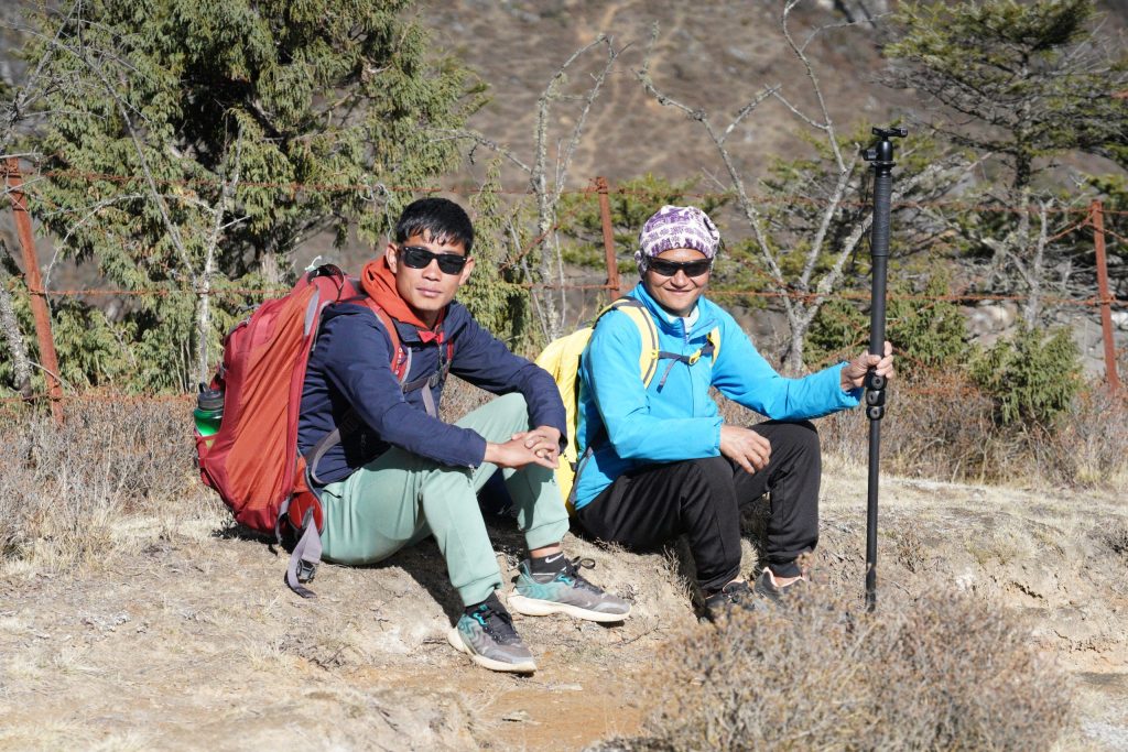 Two Sherpa guides rest beside a mountain trail in Nepal, wearing sunglasses and hiking gear after a day on the trail. One carries a large backpack while the other holds a folded tripod, with evergreen trees and the rugged Himalayan landscape in the background. Above Namche Bazaar, Nepal 2025