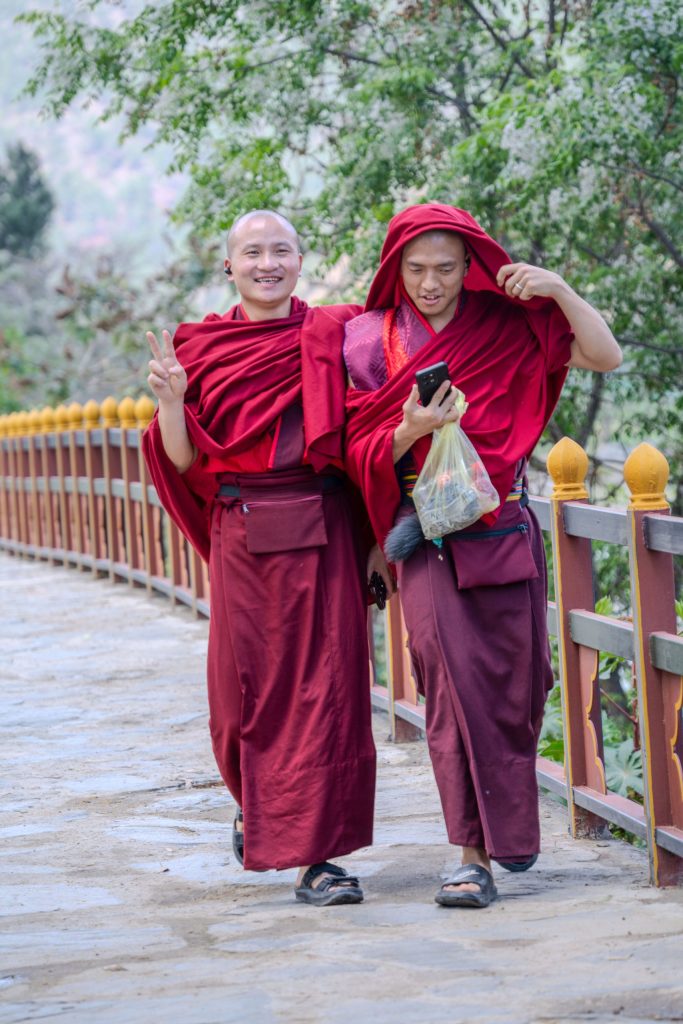 Two young Buddhist monks in crimson robes walk across a bridge in Bhutan. One smiles and flashes a peace sign toward the camera, while the other looks at a smartphone beneath his hood, blending ancient tradition with modern technology. Bhutan 2025