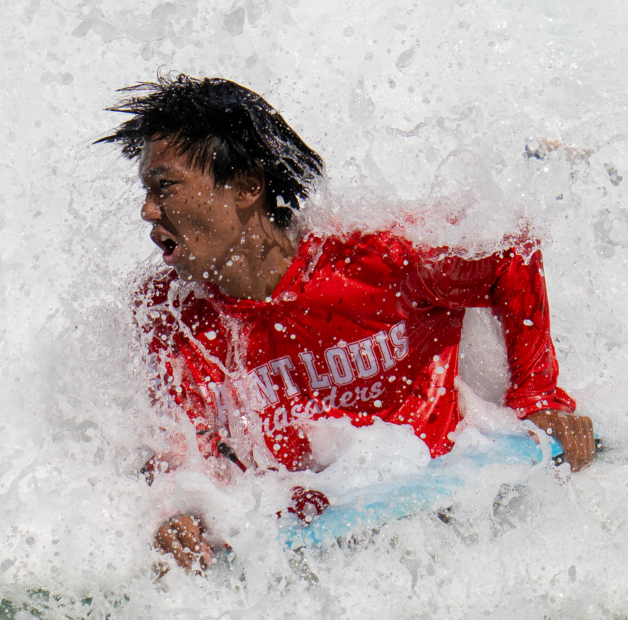 A surf student wearing a bright red rash guard is engulfed by a crashing wave while holding onto a blue surfboard on the North Shore of Oahu, Hawaii. 2026