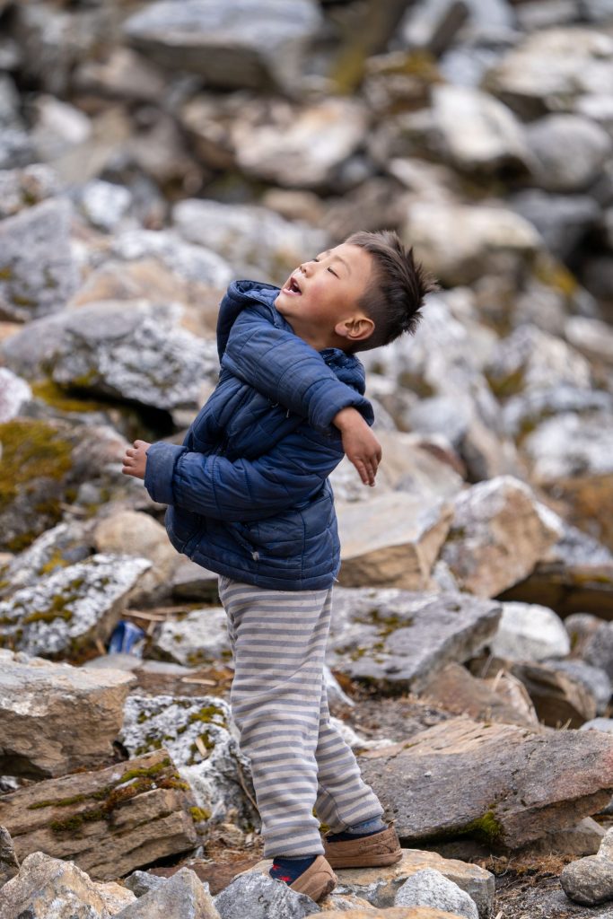 A young boy in Khumjung Village, Nepal, stands among rocky terrain with his body leaning back and his arm extended after throwing a soccer ball to a friend. His joyful expression and animated posture capture the carefree energy of childhood in the Himalayan village. Nepal 2025