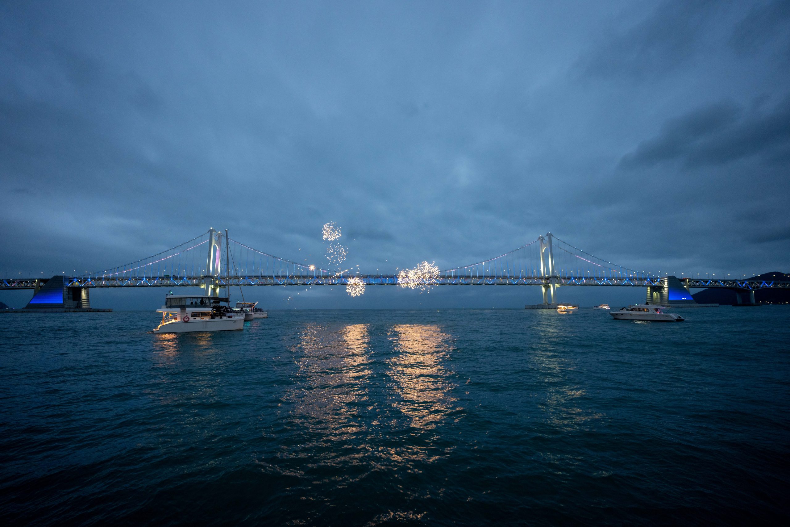 Fireworks burst above Gwangan Bridge (Diamond Bridge) at dusk, illuminating the suspension bridge and reflecting across the waters of Gwangalli Bay as boats watch from below. South Korea 2026