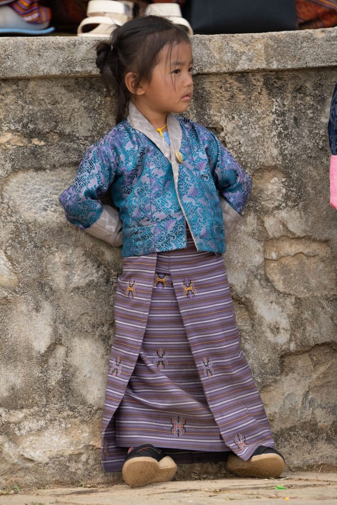 young Bhutanese girl dressed in a traditional kira and colorful silk jacket leans against a stone wall during the Paro Tshechu festival. With her hands tucked behind her back and a thoughtful expression, she watches the festivities unfold. Bhutan 2025