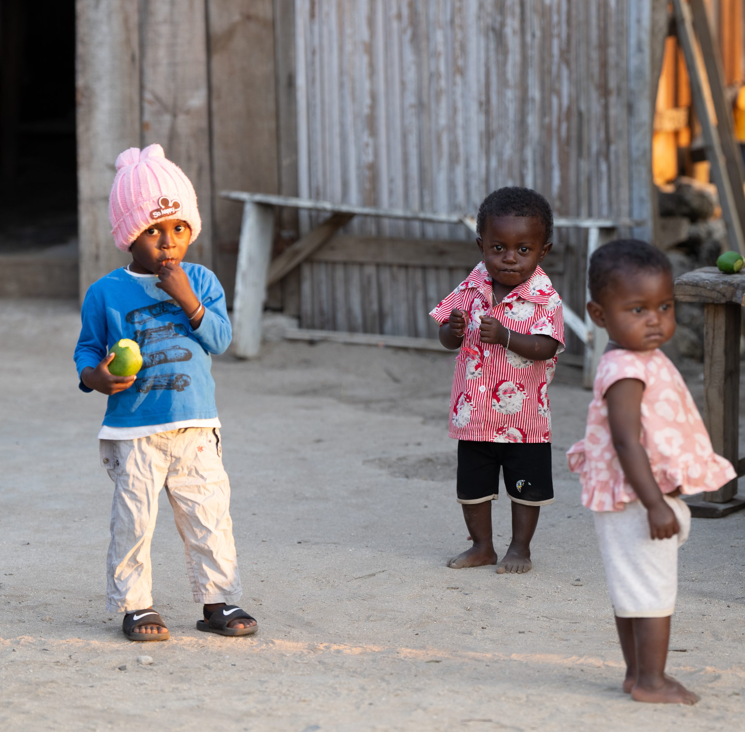 Three young children stand outside a wooden building at Miavana by Time + Tide on Nosy Ankao, Madagascar. One child holds a green mango while the others look curiously toward the camera. Madagascar 2025
