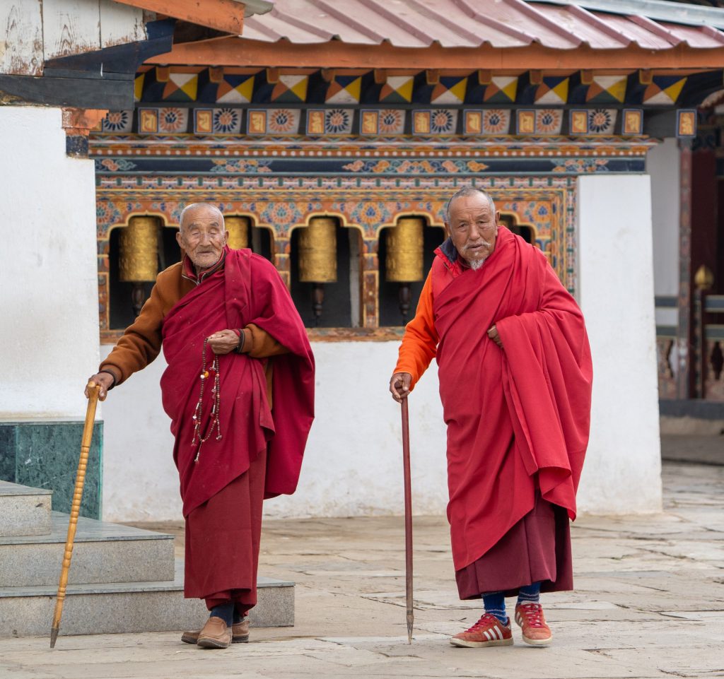 Two elderly Buddhist monks in deep red robes walk through a monastery courtyard, each using a wooden walking stick. One monk carries a string of prayer beads, while colorful prayer wheels and ornate Bhutanese architecture form the backdrop. Bhutan 2025