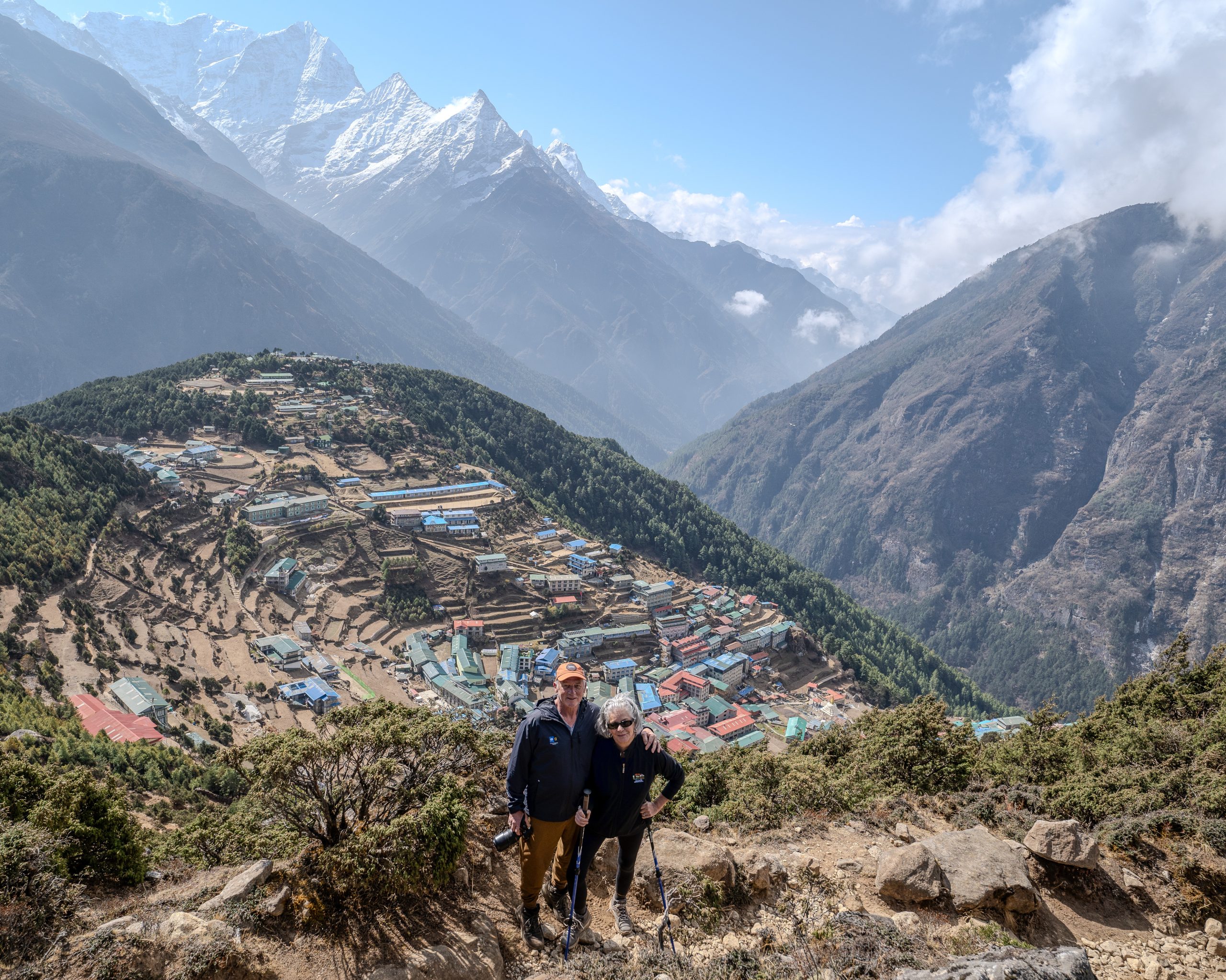 Two hikers stand on a rocky overlook above Namche Bazaar in Nepal, with the colorful mountain town below and snow-capped Himalayan peaks rising dramatically into a clear blue sky. Nepal 2025