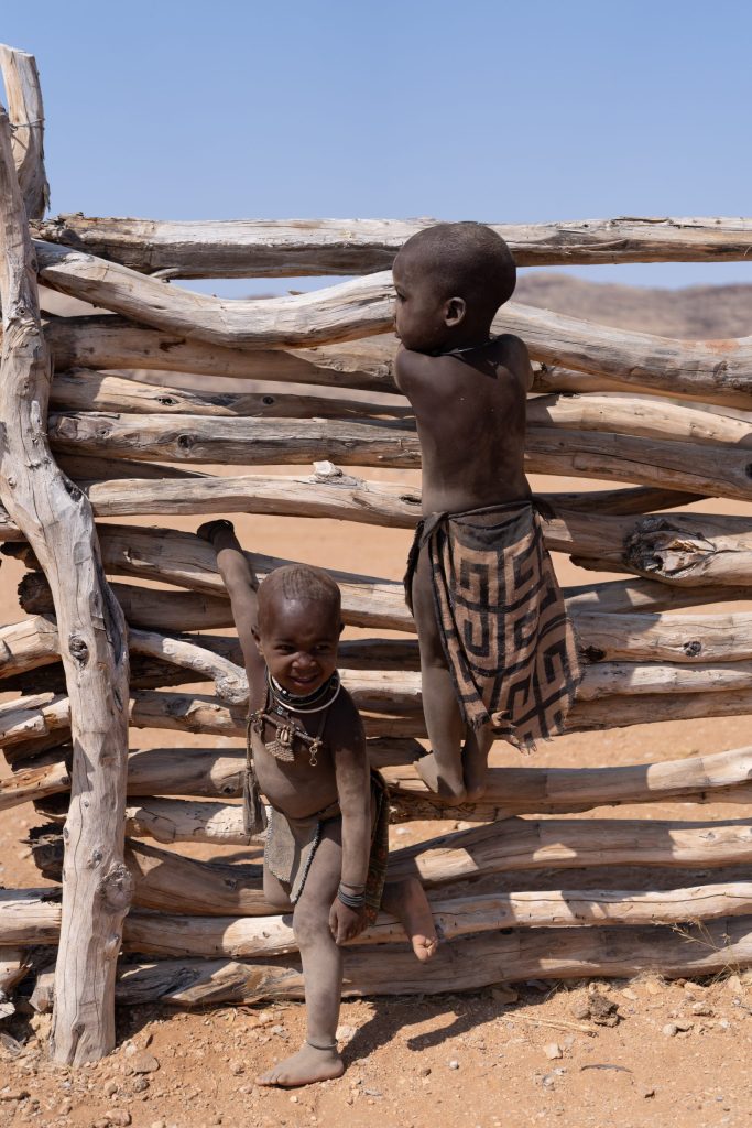 Two Himba children play on a rustic wooden fence in a village in northern Namibia. One child smiles toward the camera while the other looks across the landscape, wearing traditional clothing and jewelry. Hoanib River Valley Namibia October 2025