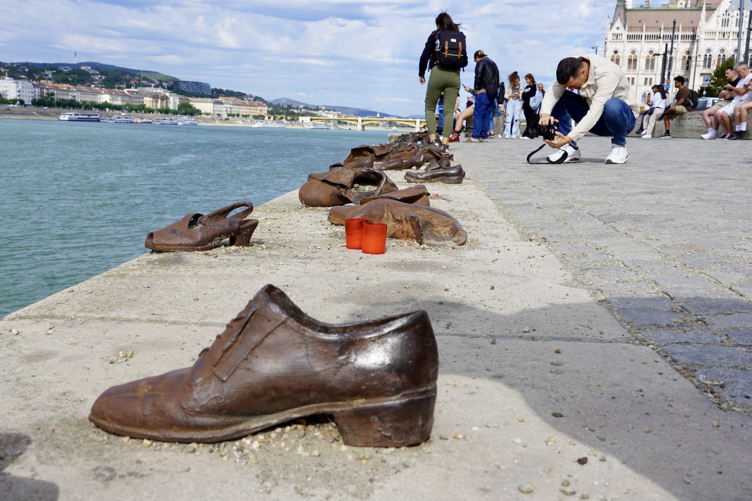 Bronze shoes from the Shoes on the Danube memorial line the riverbank in Budapest, Hungary, as visitors pause to photograph and reflect. Red memorial candles rest among the shoes, honoring the thousands of Jewish men, women, and children murdered along the Danube during World War II. Bhudapest, Hungary 2024