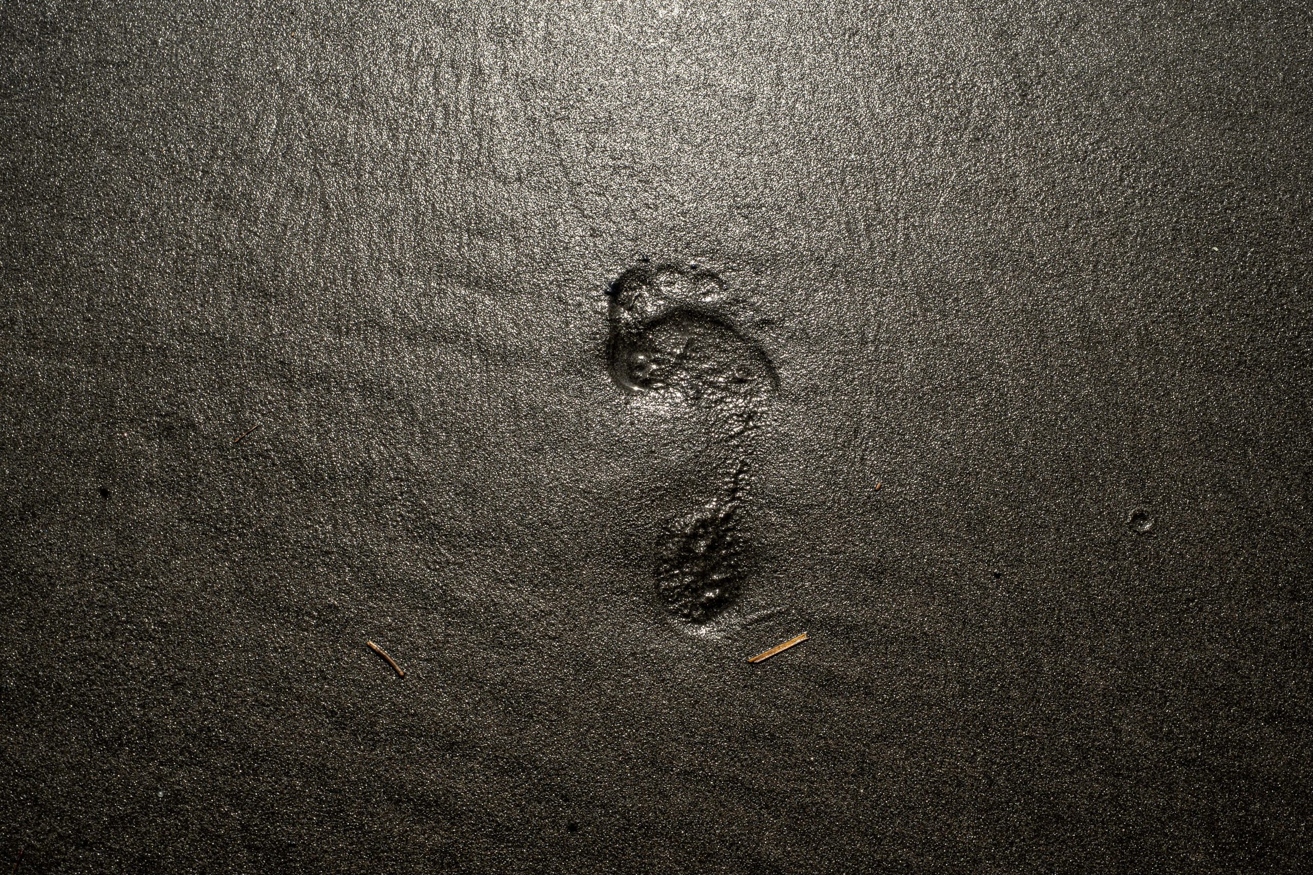 A single human footprint is pressed into smooth, dark wet sand at the point where the Tongapōrutu River meets the Tasman Sea in New Zealand. The glistening black sand reflects soft light, creating a minimalist composition that evokes solitude, movement, and the fleeting nature of our passage through the landscape.