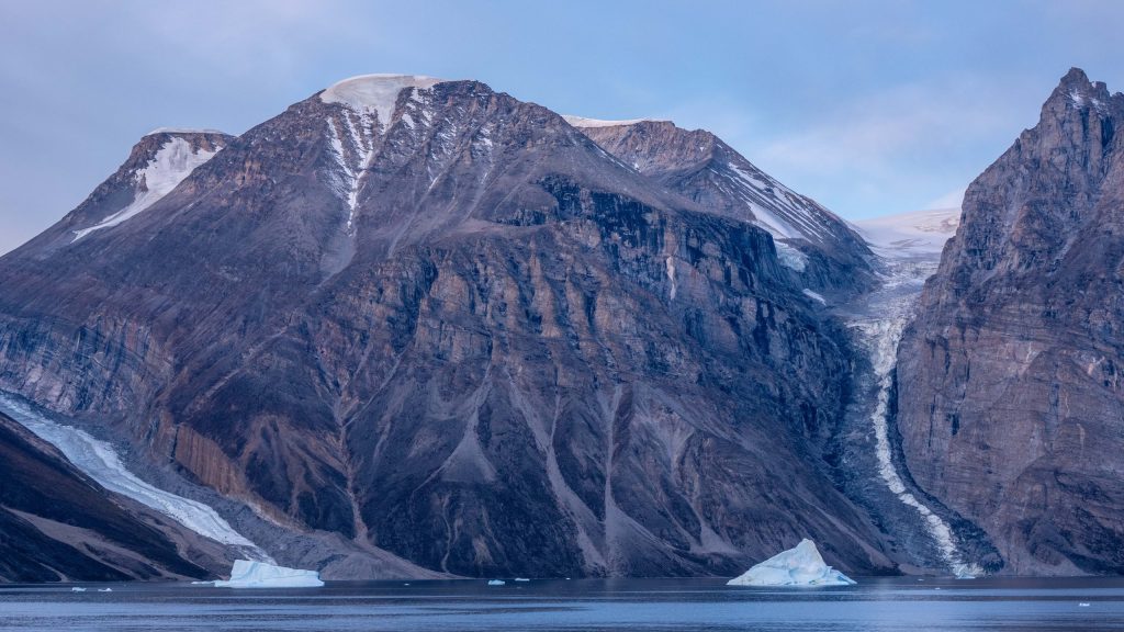 A narrow glacier descends between steep, barren mountains into a fjord in East Greenland, with exposed rock surrounding the ice and several blue-white icebergs floating in the foreground. 2026