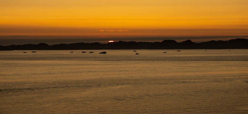 The sun sets behind dark mountain silhouettes near Nuuk, Greenland, casting deep golden-orange light across calm fjord waters dotted with small icebergs. 2026