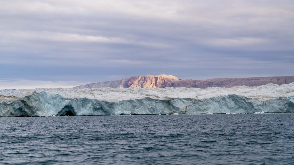 The Waltershausen Glacier stretches across a fjord in East Greenland, its blue-white ice face rising above dark Arctic water with rugged mountains illuminated by sunlight in the distance. Greenland 2026