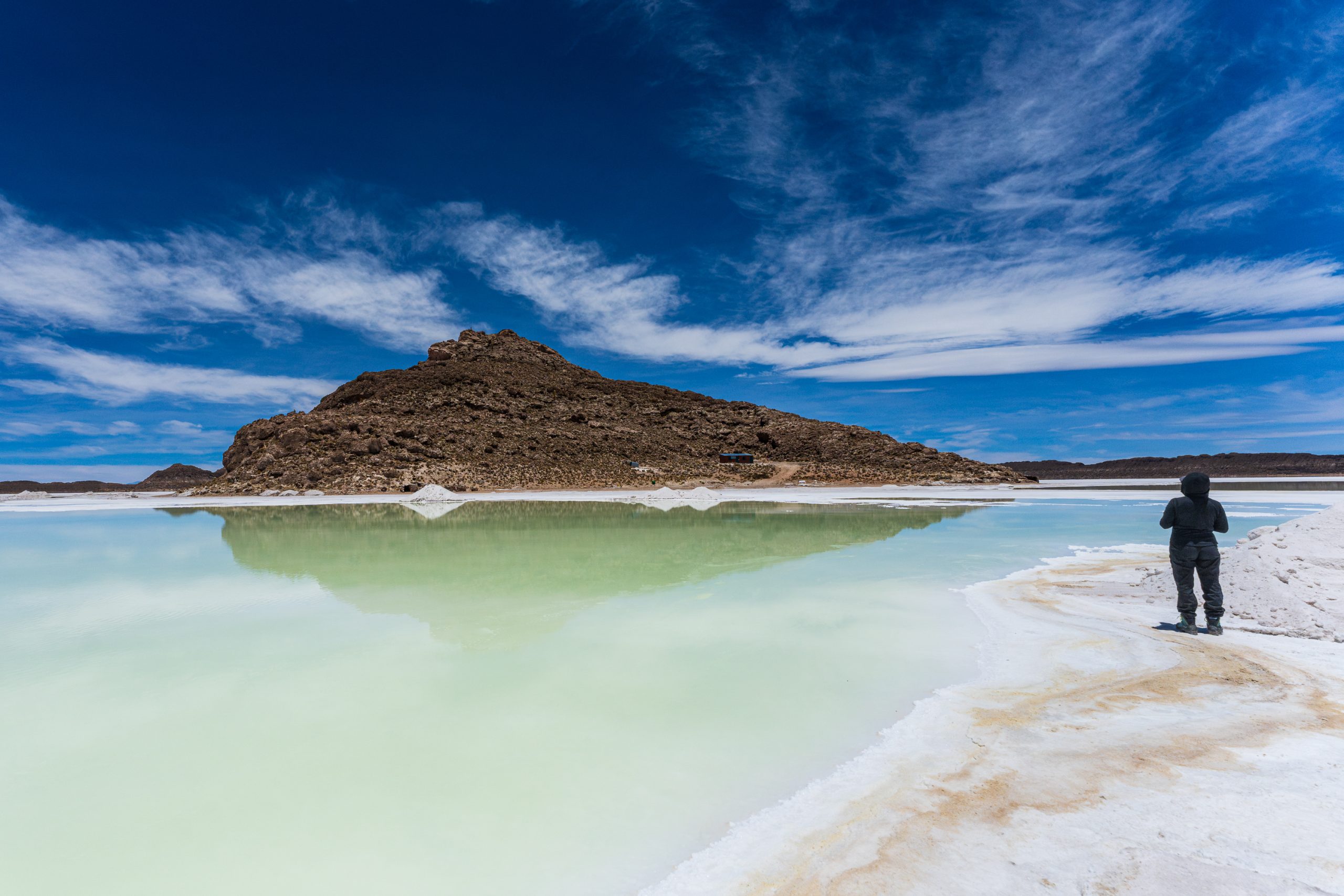 A traveler stands beside a pale turquoise saltwater lagoon reflecting a rocky island beneath a brilliant blue sky with wispy clouds in Bolivia. 2026