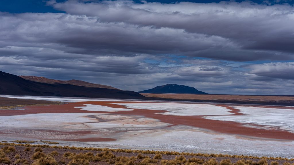 Sweeping red and white mineral patterns across a high-altitude lagoon in Bolivia's Altiplano, beneath dark volcanic mountains and dramatic storm clouds. 2026