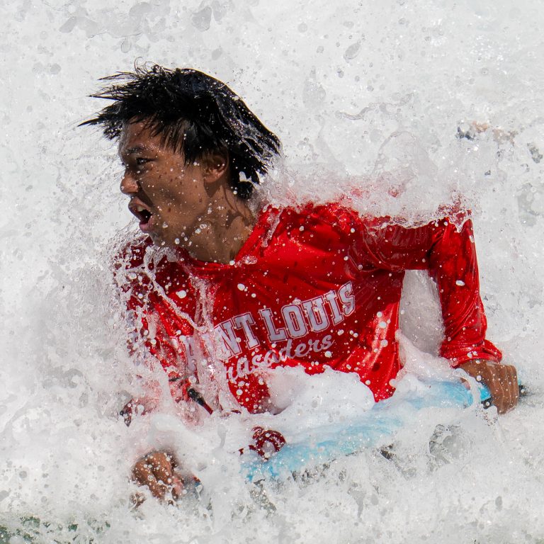 A surf student wearing a bright red rash guard is engulfed by a crashing wave while holding onto a blue surfboard on the North Shore of Oahu, Hawaii. 2026