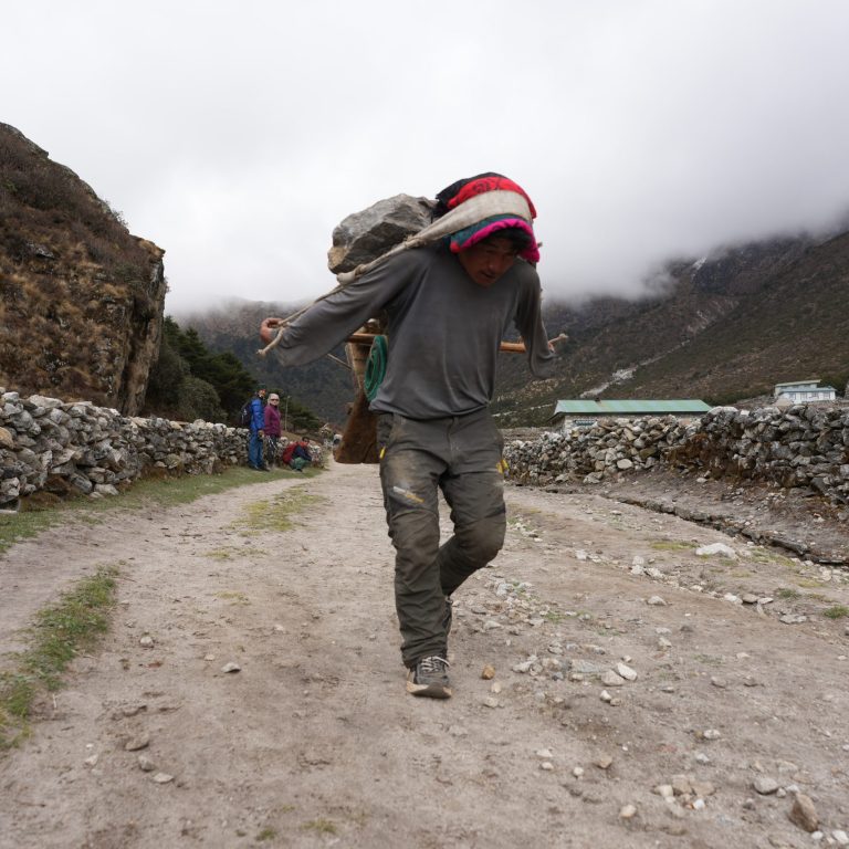 A Sherpa laborer carries a large load of rocks in a woven back basket supported by a forehead strap as he walks along a dirt trail in the Everest region of Nepal. Stone walls line the path, while low clouds cling to the surrounding Himalayan mountains, highlighting the demanding conditions in which Sherpas build strength and endurance. Namche Bazaar, Nepal 2025