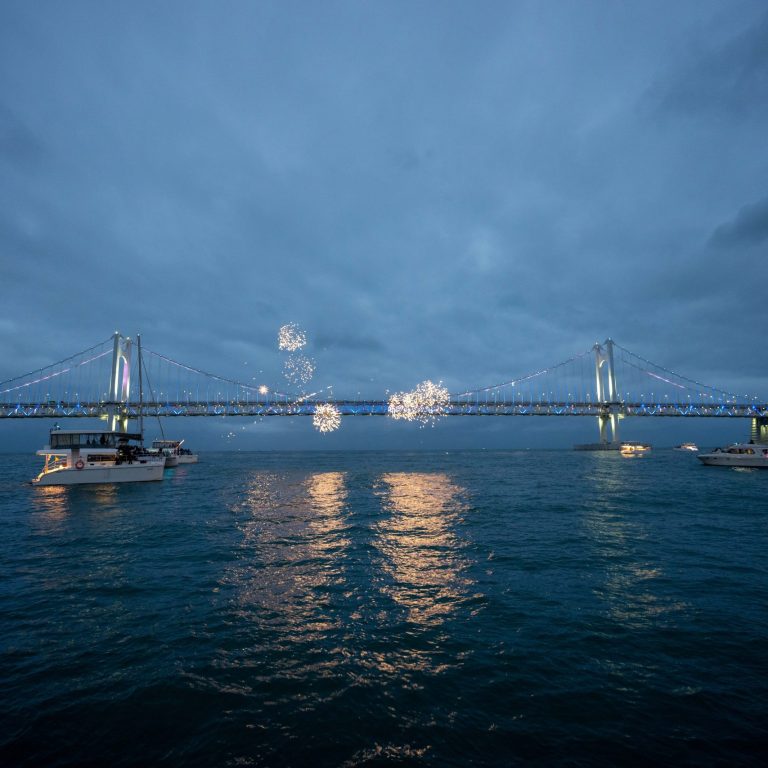 Fireworks burst above Gwangan Bridge (Diamond Bridge) at dusk, illuminating the suspension bridge and reflecting across the waters of Gwangalli Bay as boats watch from below. South Korea 2026