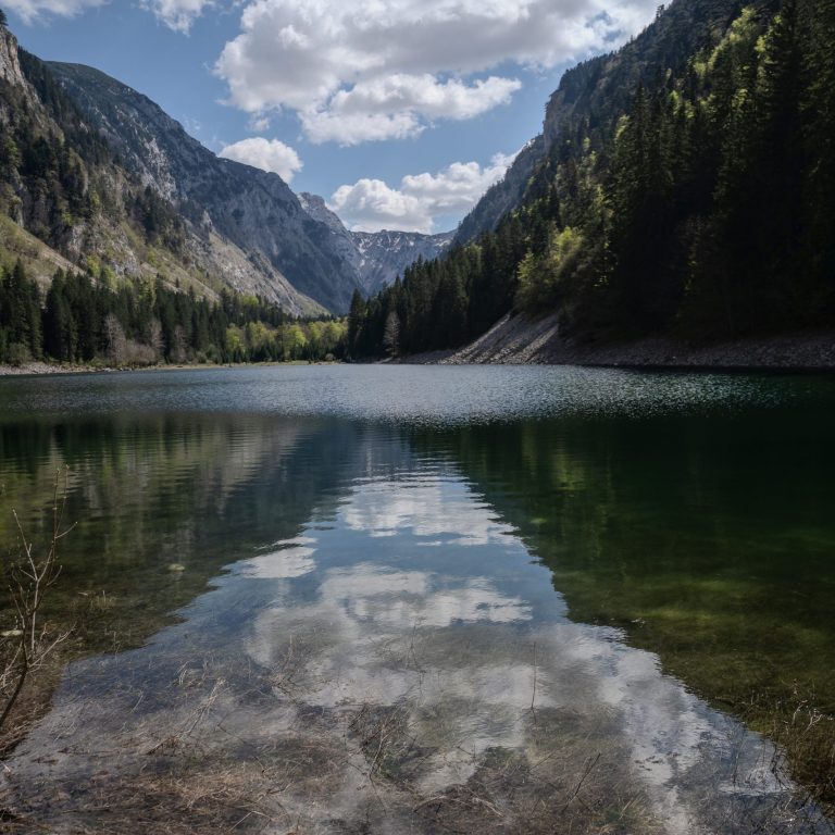 A crystal-clear mountain lake in Durmitor National Park, Montenegro, reflects towering forested slopes, rugged limestone peaks, and billowing white clouds beneath a bright blue sky. 2024